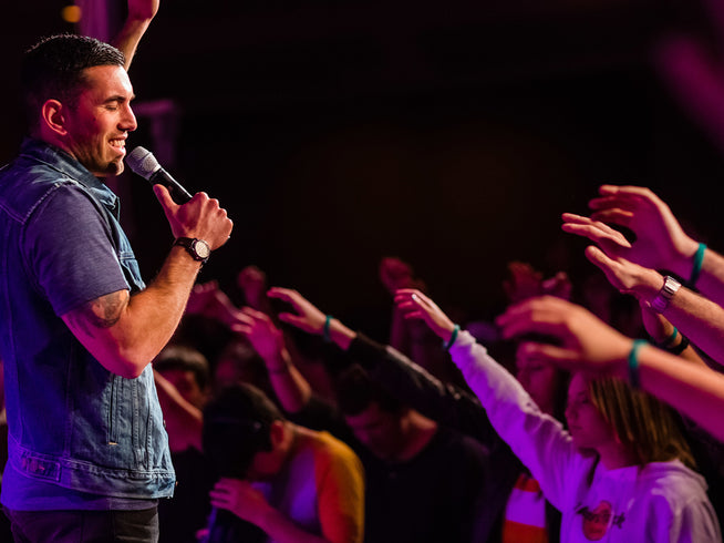 Man holding a microphone in front of an audience with raised hands, likely at a concert or event.