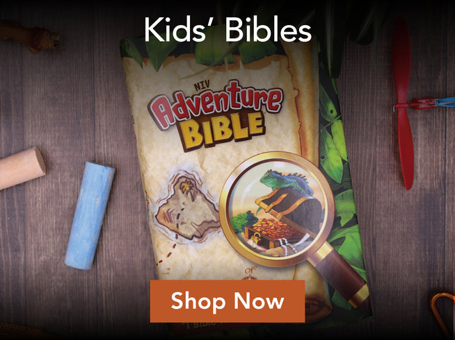 A children's Bible titled 'New Adventure Bible' displayed on a dark wooden surface with props like chalk, a magnifying glass illustration, and a red propeller. White text at the top reads 'Kids' Bibles' and an orange button at the bottom says 'Shop Now'.