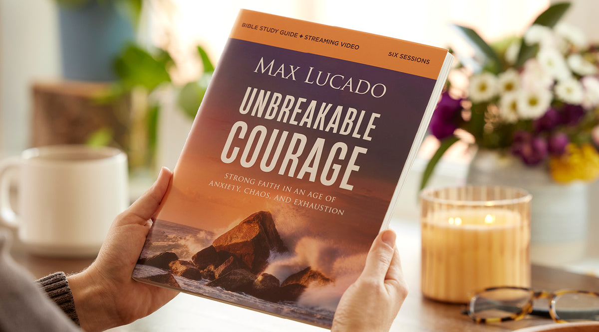 A person holds a copy of the book 'Unbreakable Courage' by Max Lucado in a cozy, softly lit indoor setting with a mug, candle, and flowers in the background.