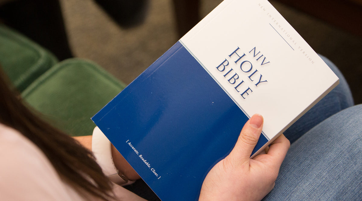 A person holding a blue and white 'NIV Holy Bible' in their lap, with their hand visible and jeans in the background.