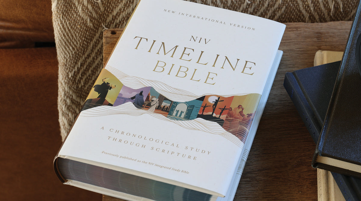 A hardcover copy of the NIV Timeline Bible lies open on a wooden table, with other books partially visible nearby.
