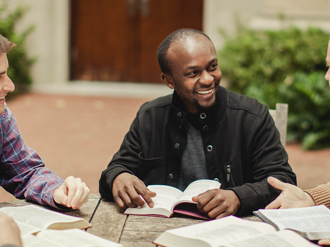 Four students are sitting outdoors, engaged in a discussion with books open on a table.