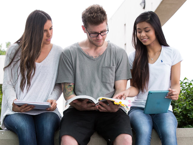 Three students sitting outdoors, reading books and using a tablet.