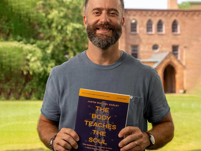 Man holding a book titled 'The Body Teaches the Soul' outdoors with a building in the background