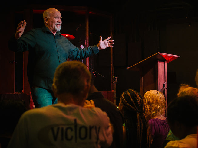 Man on stage with outstretched arms addressing an audience, with a person wearing a 'Victory' shirt in the foreground.
