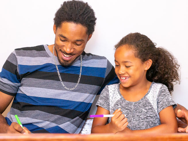 Man and young girl sitting together, smiling and holding pens, likely engaged in a creative activity.