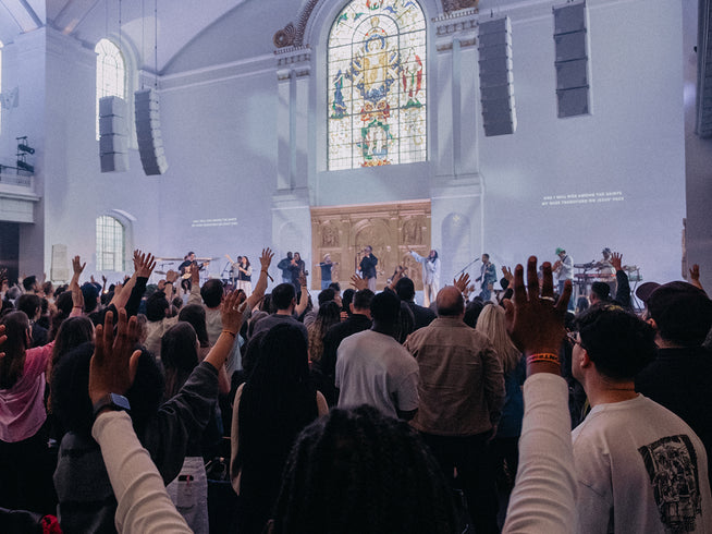 A congregation raising their hands in worship inside a church, with a worship band performing on stage in front of a large stained glass window.