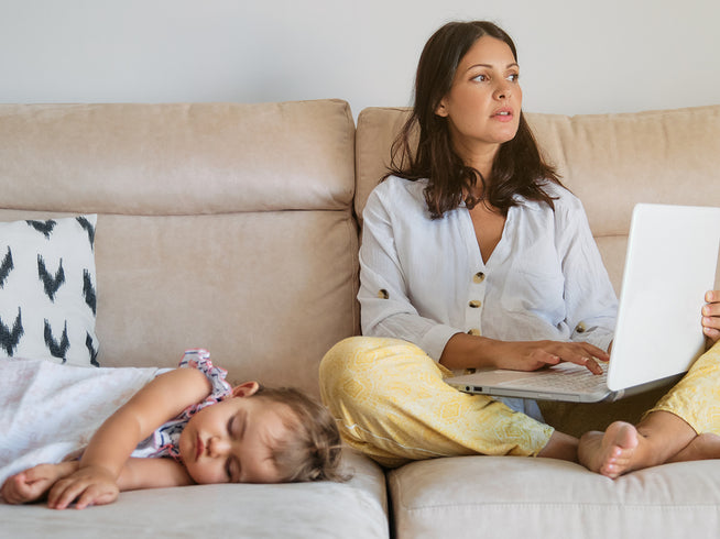 Woman sitting on a couch working on a laptop while a child rests nearby
