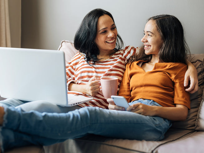 Mom and daughter sitting on a couch talking to each other. One has a cup of coffee and a laptop, the other has a mobile device.