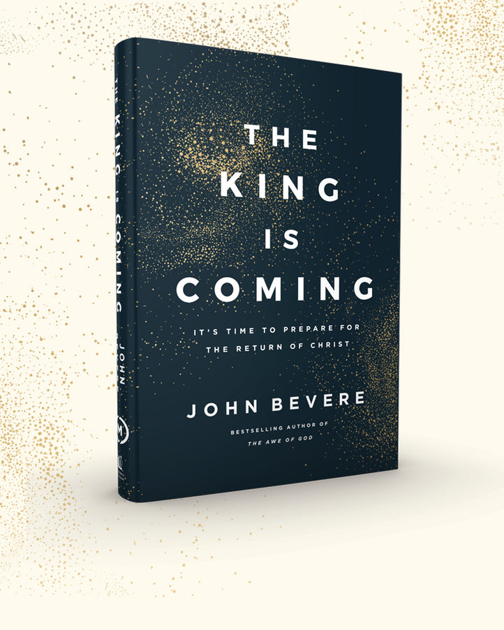 The King Is Coming book by John Bevere, featuring a dark cover with gold glitter speckles and white text, shown standing upright on a light cream background with gold speckles.